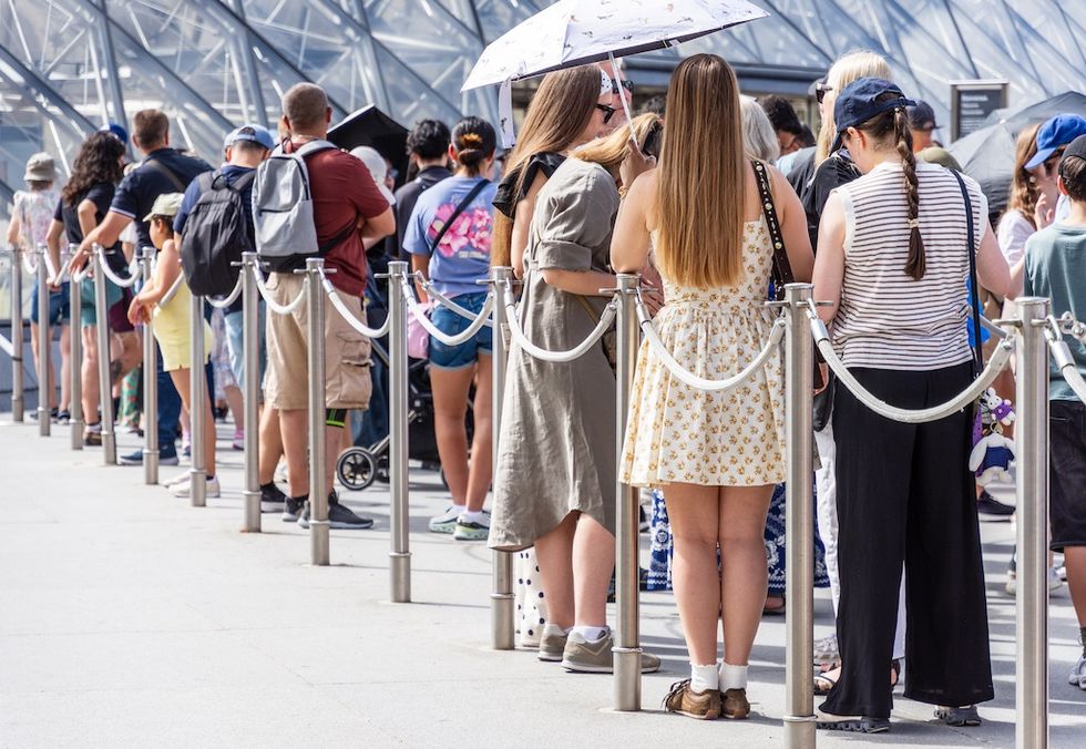 People in line outside a modern building, some using umbrellas for shade.