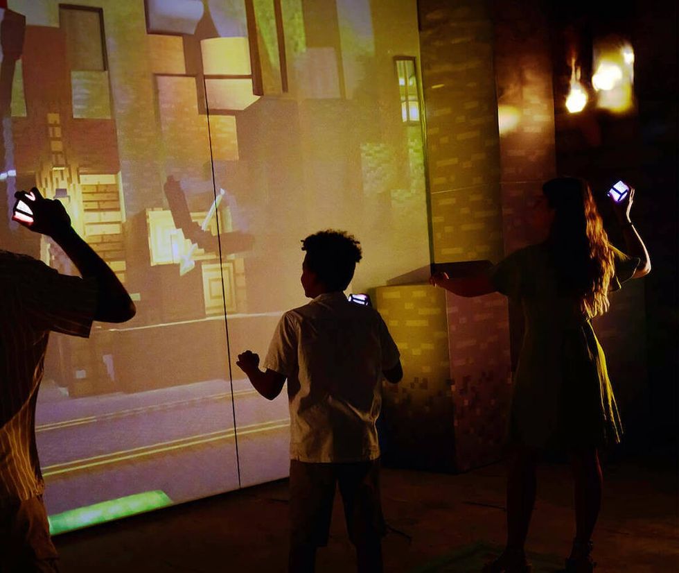 People interact with a digital wall projection using handheld devices during Minecraft Experience: Villager Rescue