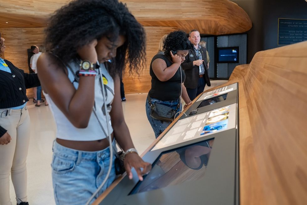 People interact with a touchscreen exhibit in a modern, wood-paneled museum space.