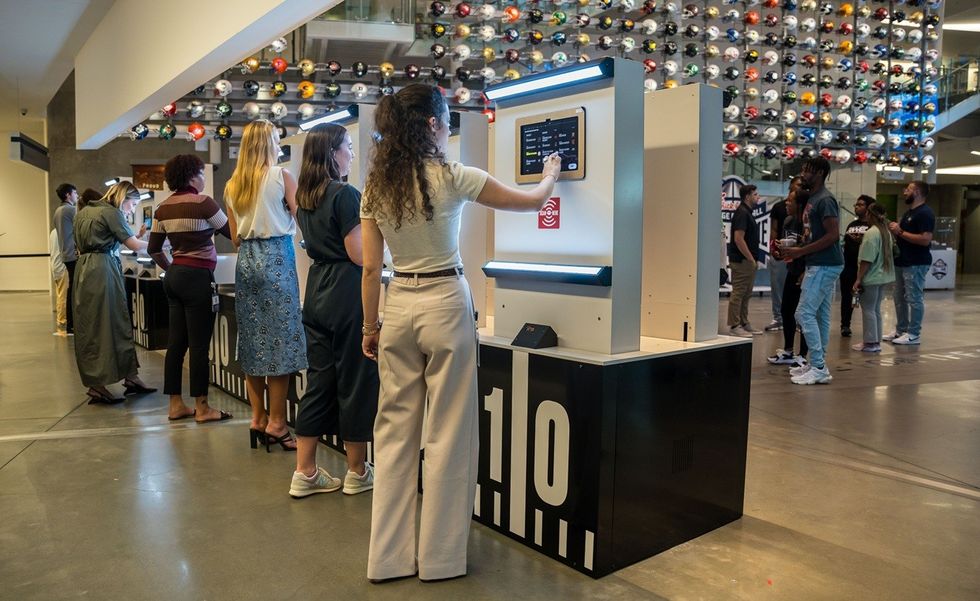 People interacting with kiosks in a modern building with a wall of helmets.