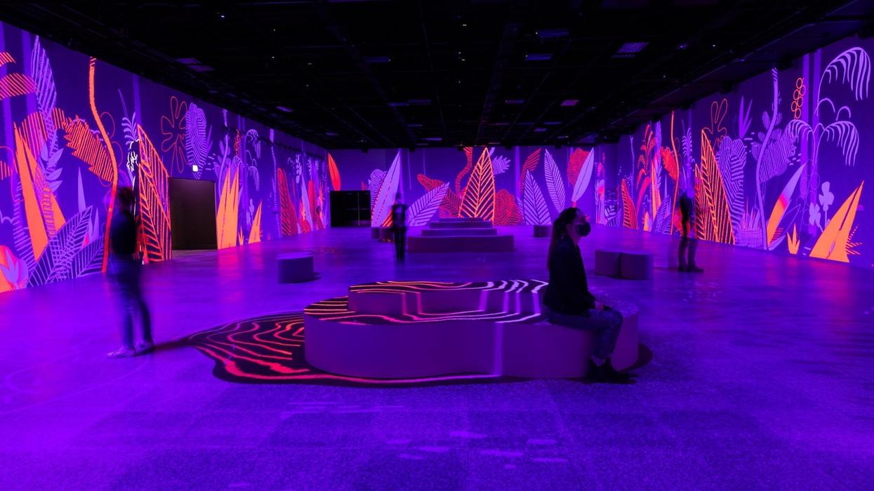 People observe vibrant neon floral art projected in a dark gallery space.