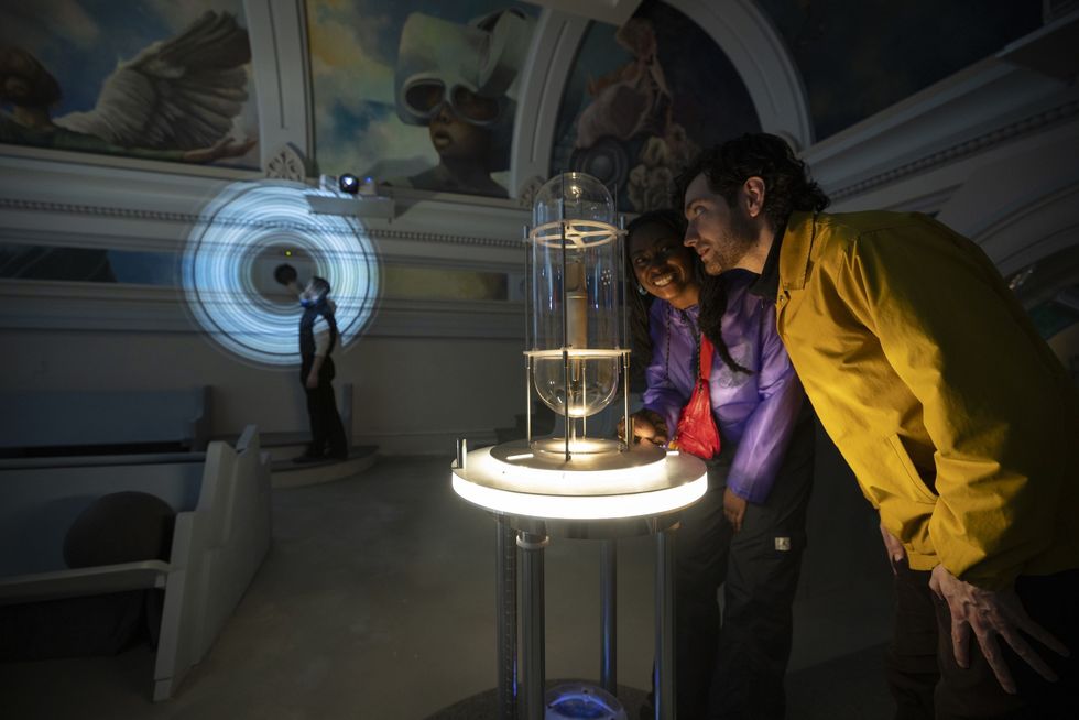 People observing a glowing scientific glass installation in a dimly lit room with murals at the Ministry of Awe.