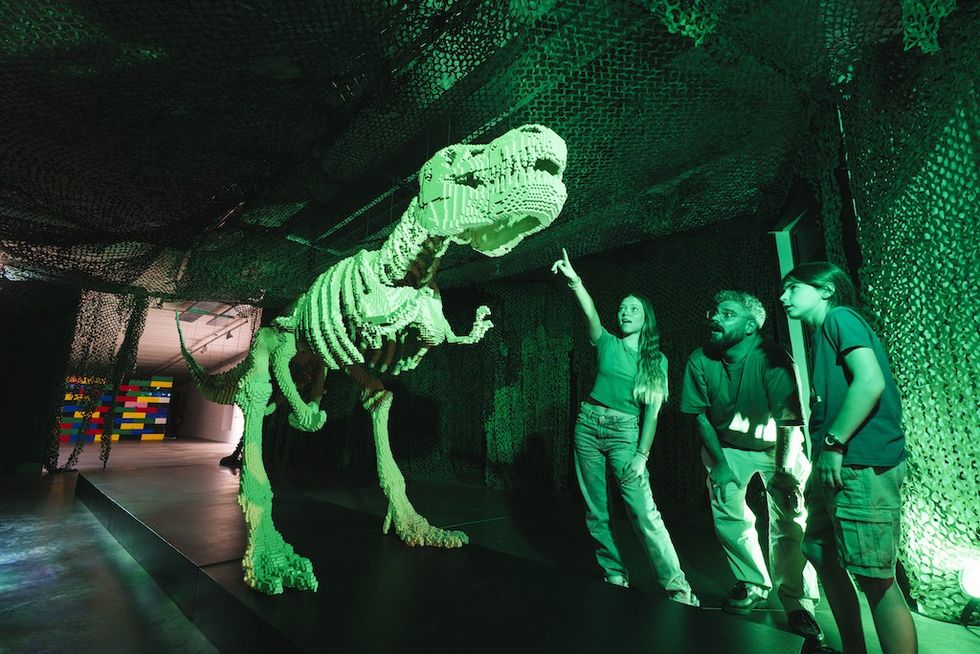 People observing a green-lit Lego dinosaur sculpture in an exhibit room.