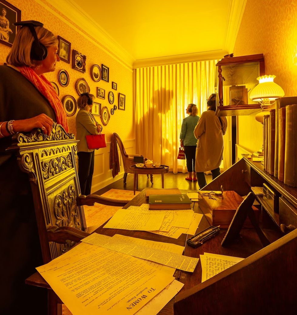 People observing a room display with vintage decor and a shadowed figure behind yellow curtains. The War Rooms at The Story of Emily