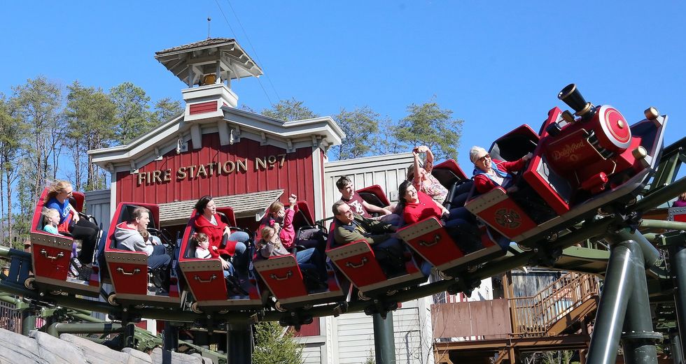People on a red roller coaster near a fire station-themed building.