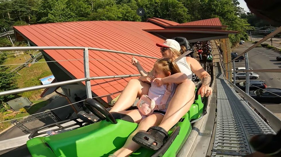 People on a roller coaster, surrounded by trees, riding next to a red-roofed building.