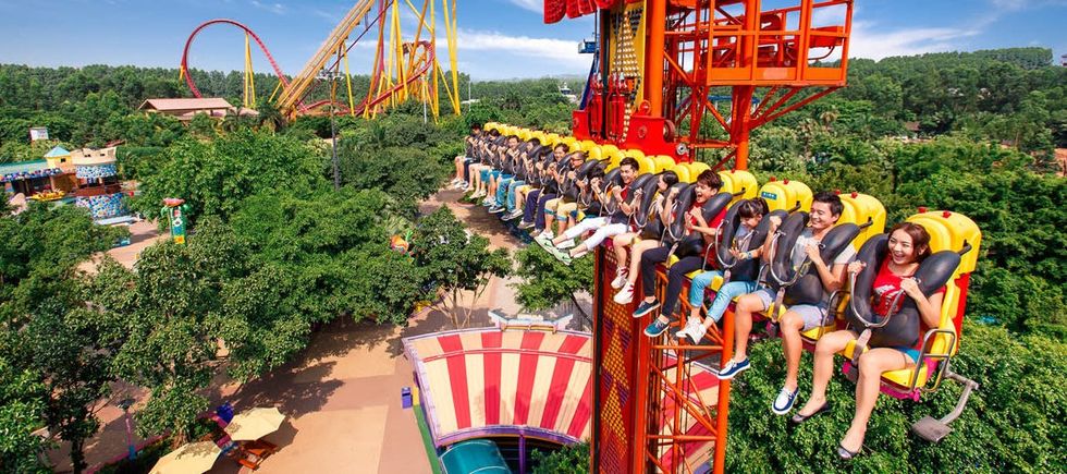 People on a thrill ride ascending in an amusement park called Chimelong Paradise surrounded by trees and roller coasters.