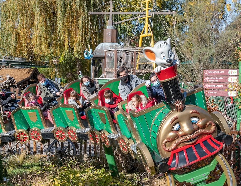 People on a whimsical train-themed roller coaster ride with a mouse mascot, surrounded by trees.