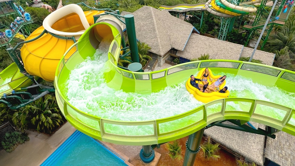 People on a yellow raft sliding down a curvy green water ride named PowerRAPIDS at Aquatopia Water Park in Vietnam.