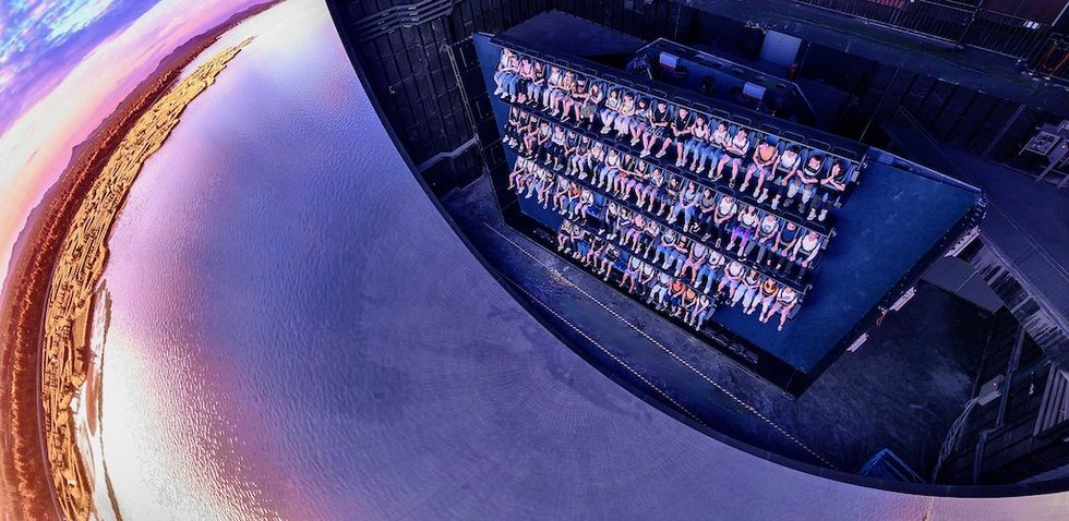 People on an indoor ride experience, facing a projected curved simulation of a landscape.