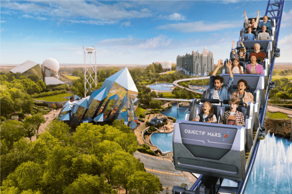 People on Objectif Mars roller coaster at Futuroscope amusement park with futuristic buildings in the background.