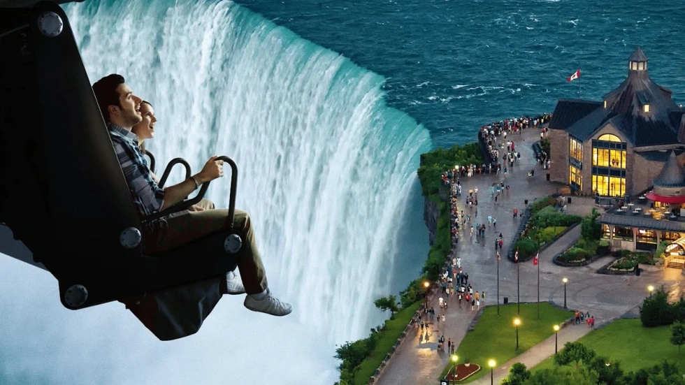 People on ride above a scenic waterfall, overlooking a lit-up building and pathway below.