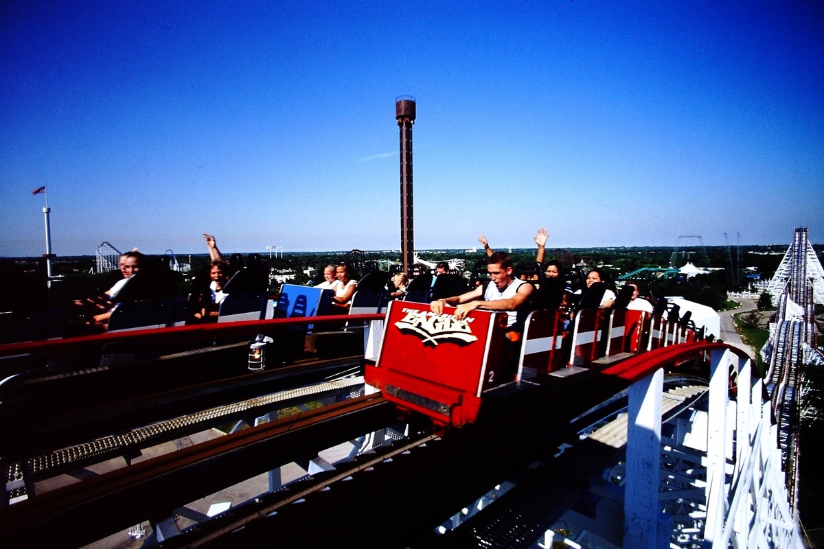 People on the red and blue American Eagle roller coaster at Six Flags Great America, arms raised, against a clear blue sky.