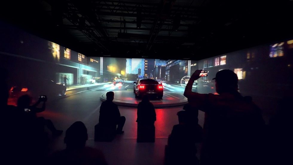 People photographing a car in a room with vibrant cityscape projections.