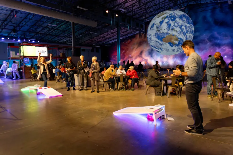 People playing indoor cornhole in a space-themed venue with a mural of Earth.