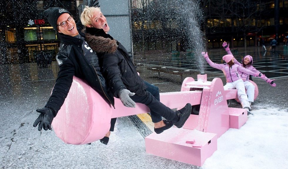 People playing on a pink seesaw that sprays snow, surrounded by snow and wearing winter clothes.