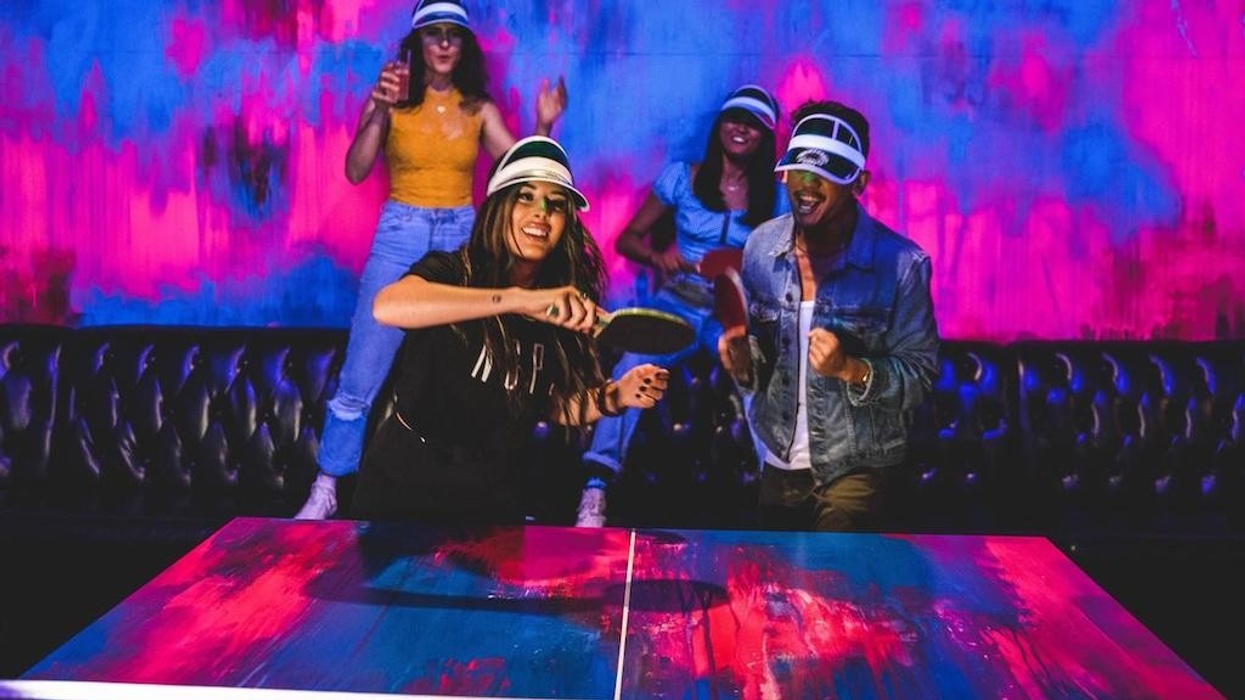 People playing ping pong in neon-lit room, wearing visors and colorful attire.