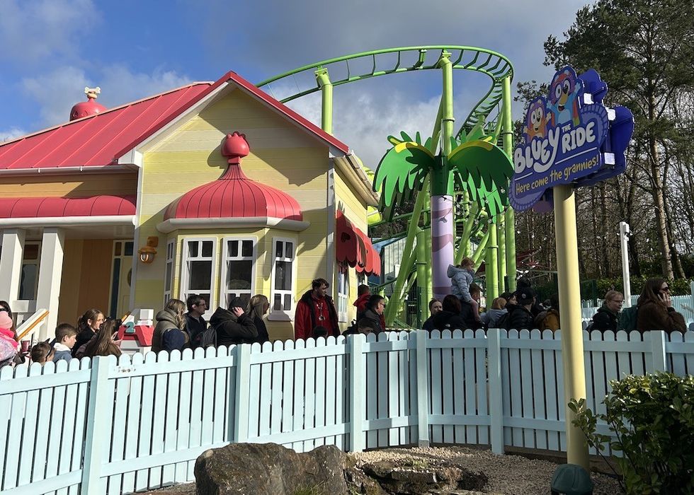 People queue outside cartoon-themed ride with a colorful house and green roller coaster.