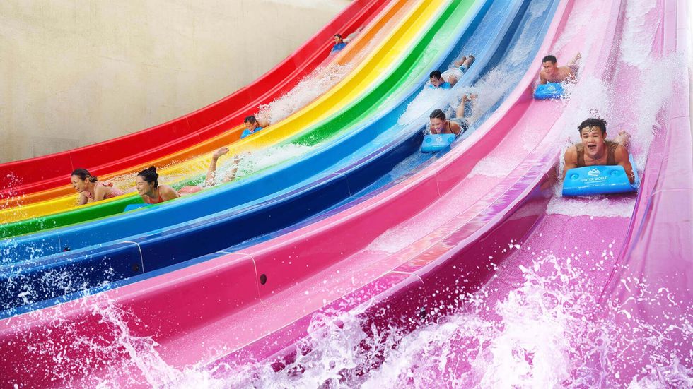 People racing down vibrant, multi-colored water slides on mats at Water World Ocean Park, splashes of water around.