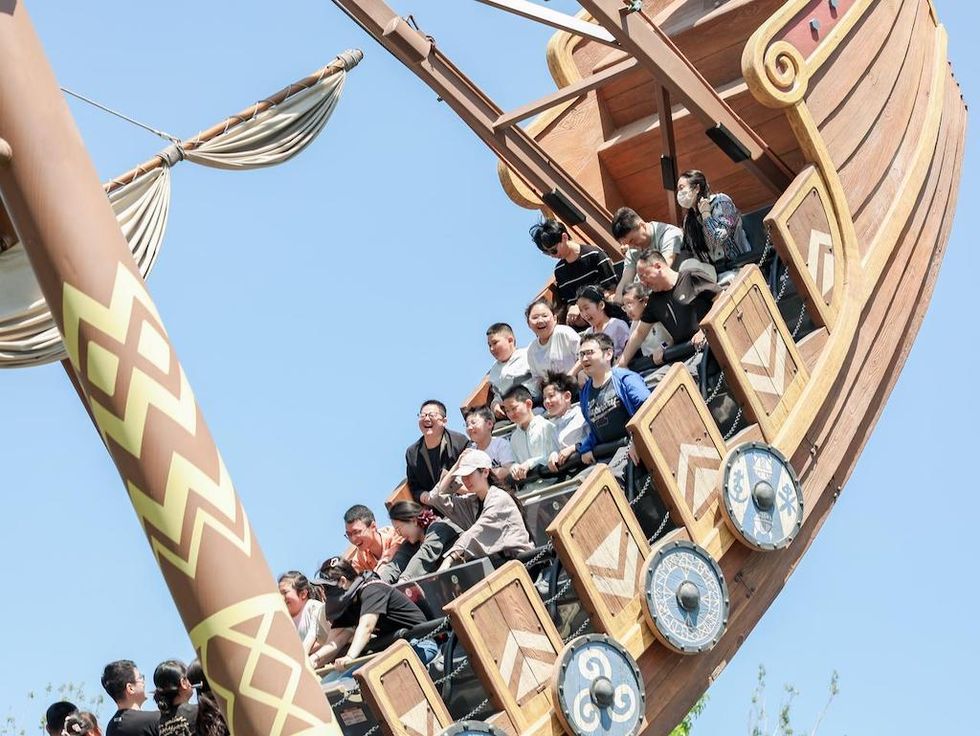 People ride a large wooden pirate ship swing at an amusement park under a clear blue sky.