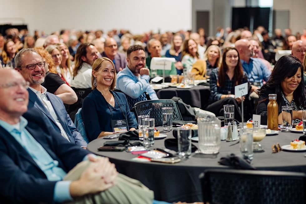 People seated at round tables, smiling and attentively watching an event, indoors.