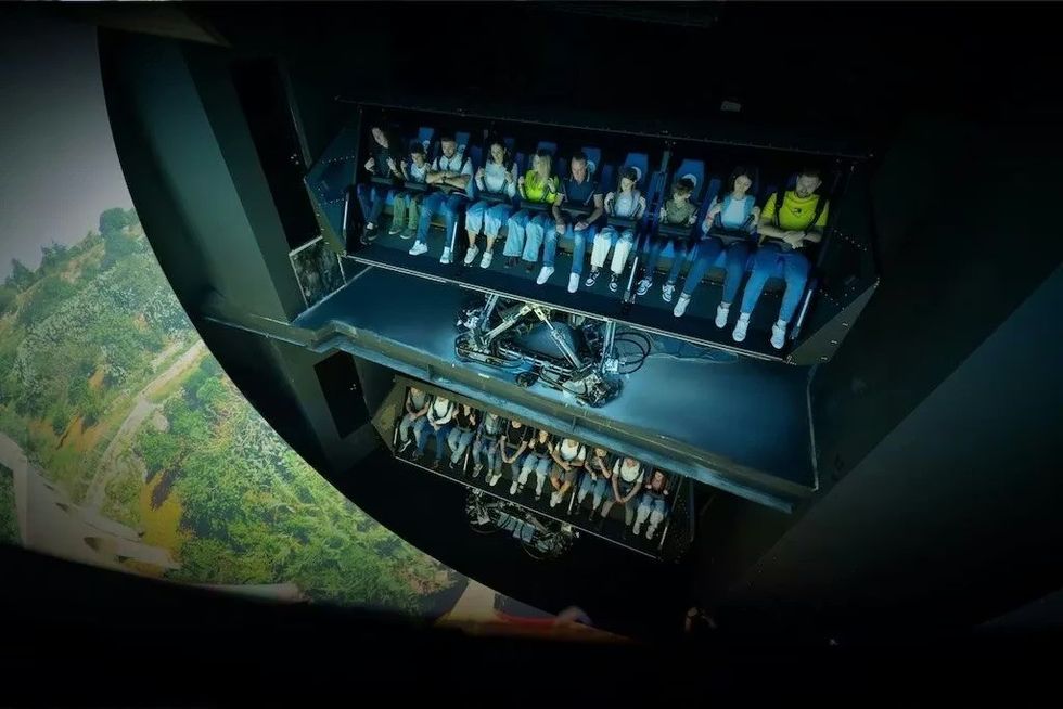 People seated in a motion simulator ride above a large screen displaying landscapes.