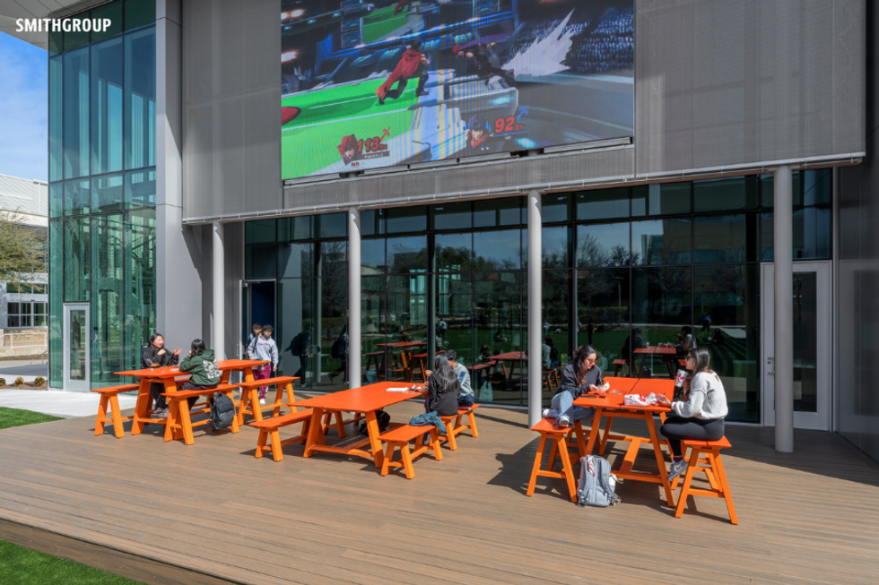 People sit at orange tables outside a building with a large screen displaying a video game.