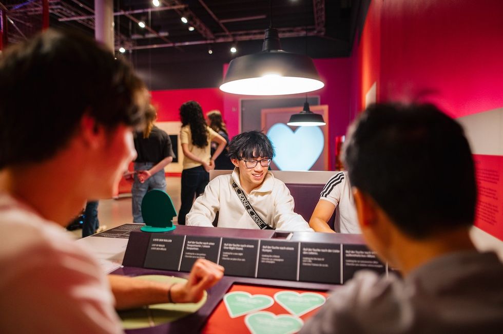 People sitting and talking at a table in a brightly lit room with pink walls during About Love exhibition, Universum Bremen