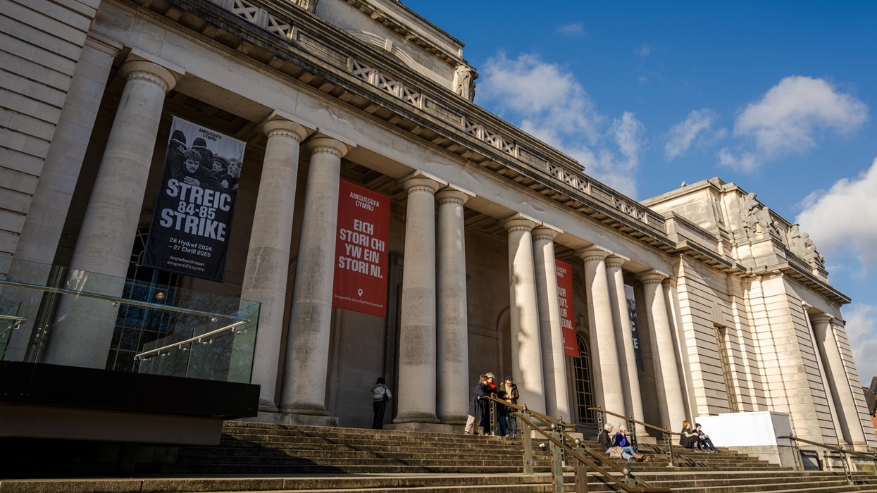 People sitting on steps outside National Museum Cardiff, a large building with columns and banners.
