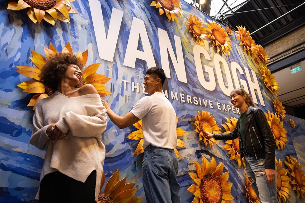 People smiling near "Van Gogh" sign with sunflowers on a blue mural at Van Gogh: The Immersive Experience in Cincinnati, US.