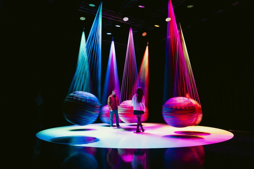 People stand on a colorful stage with large, glowing, suspended spheres in various colors.