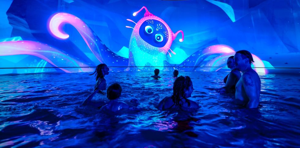 People swimming under a vibrant octopus-cat projection in a dark, neon-lit pool.