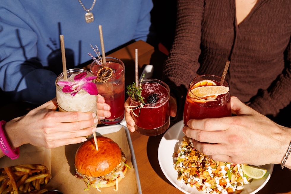 People toasting colorful cocktails over a table with burgers and fries.