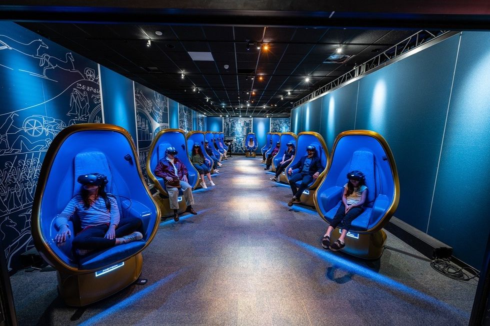 People using VR headsets in futuristic blue chairs, lined up in a dimly lit room.