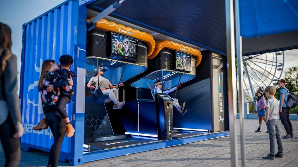 People using VR paragliding simulators inside a blue shipping container at an outdoor event.