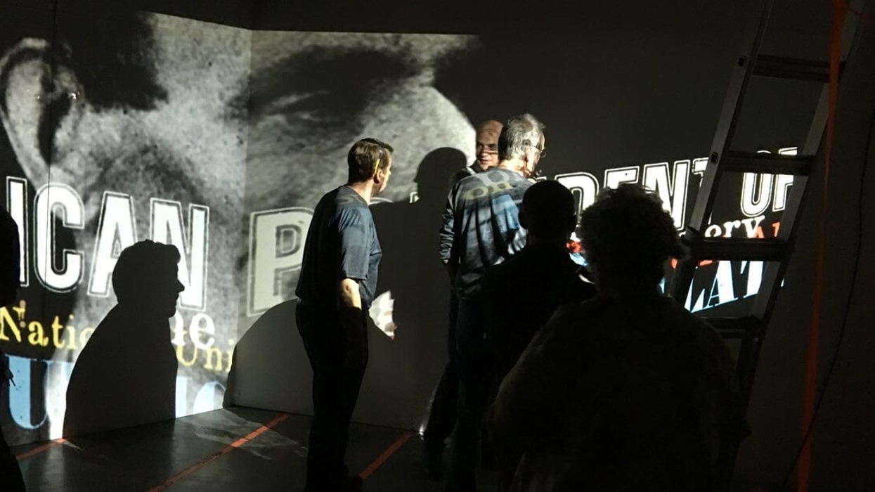 People viewing projected text and image on a dimly lit wall with a ladder on the side.
