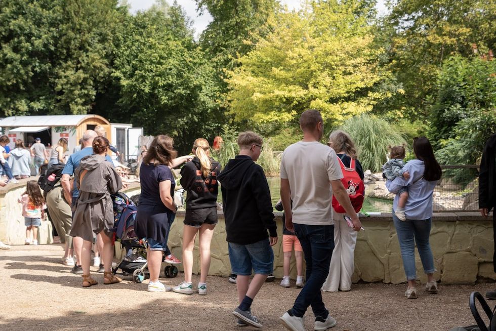 People walking and gathering outdoors at Twycross Zoo with green foliage and a water habitat in the background.