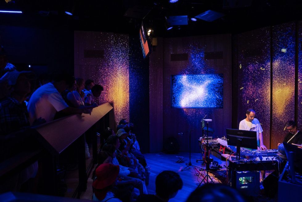 People watching musicians perform on a dimly lit stage with vibrant galaxy visuals.