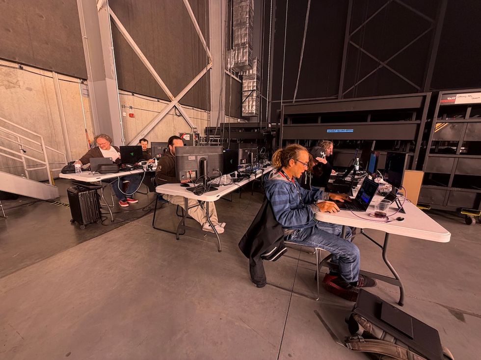 People working on laptops and computers at tables in an industrial space.