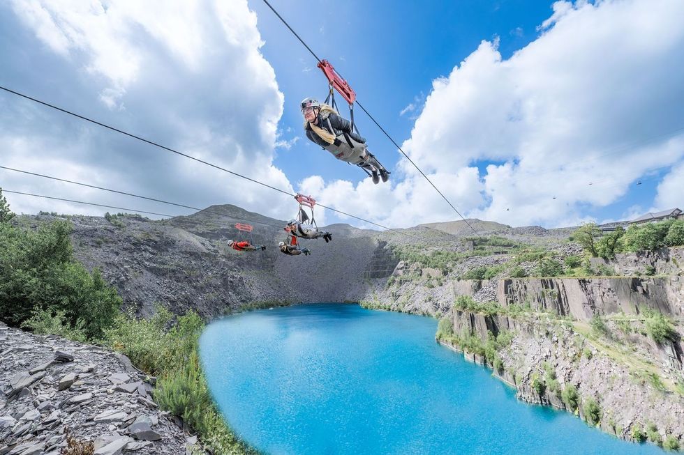 People zip-lining over a vibrant blue lake with hills and clouds in the background, Velocity ride at Zip World