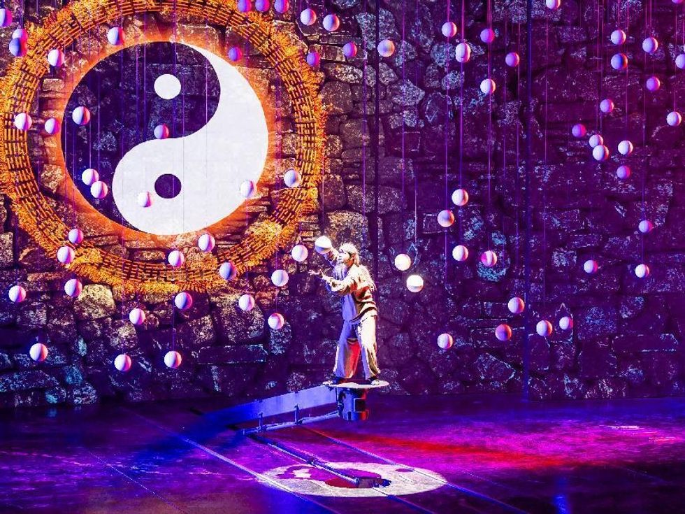 Performer balancing on beam, surrounded by balls, with large yin-yang symbol in background.