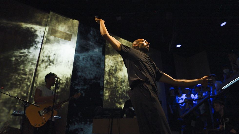Performer dances dramatically on stage with guitar player and audience in background.
