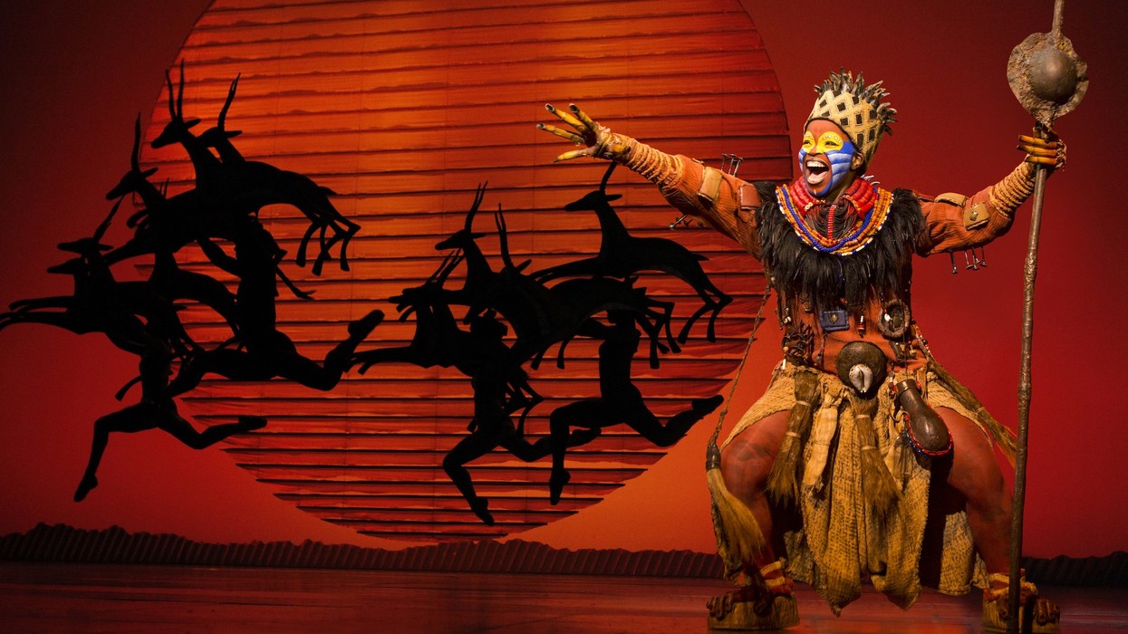 Performer in colorful costume on stage with antelope silhouettes and sunset backdrop.