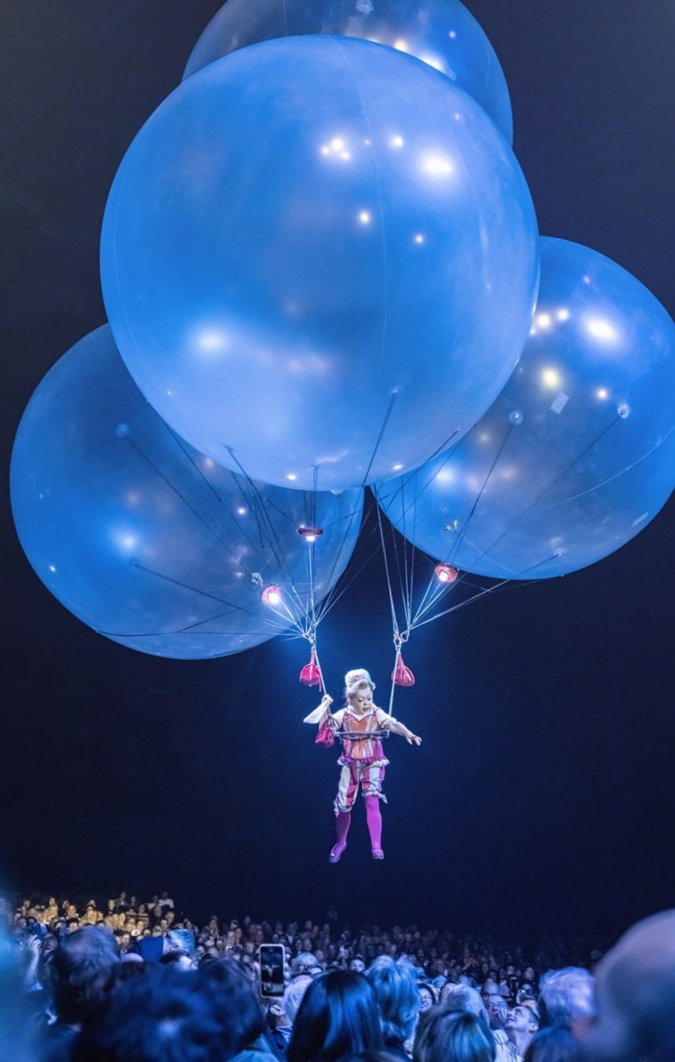 Performer in costume floats above audience with large blue balloons during Corteo by Cirque du Soleil