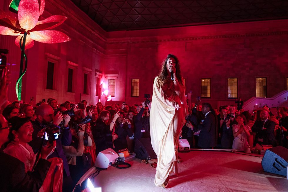Performer in flowing dress sings on stage, surrounded by a crowd under pink lighting at the British Museum
