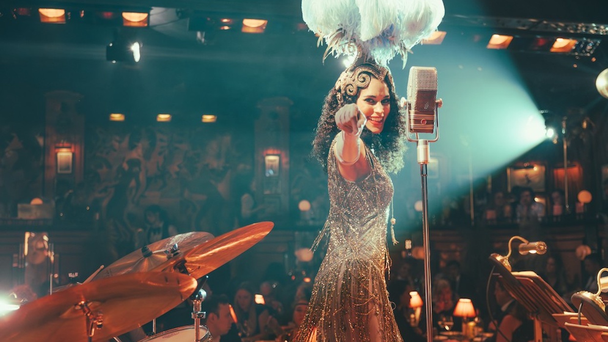 Performer in ornate dress and feathered headdress on stage with microphone and drums.