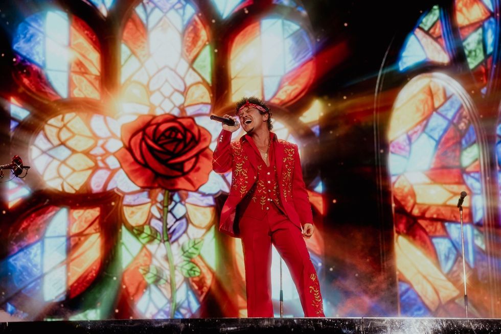 Performer in red suit sings passionately in front of vibrant stained glass backdrop.