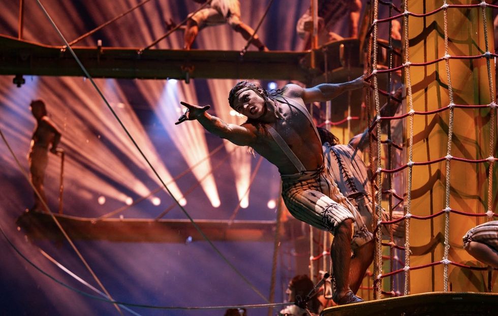 Performer in striped outfit climbs rope net in dramatic stage lighting in The House of Dancing Water.