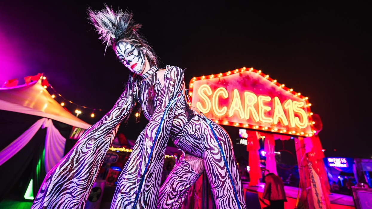 Performer in zebra print costume stands by neon "SCARE" sign at night event at Area15