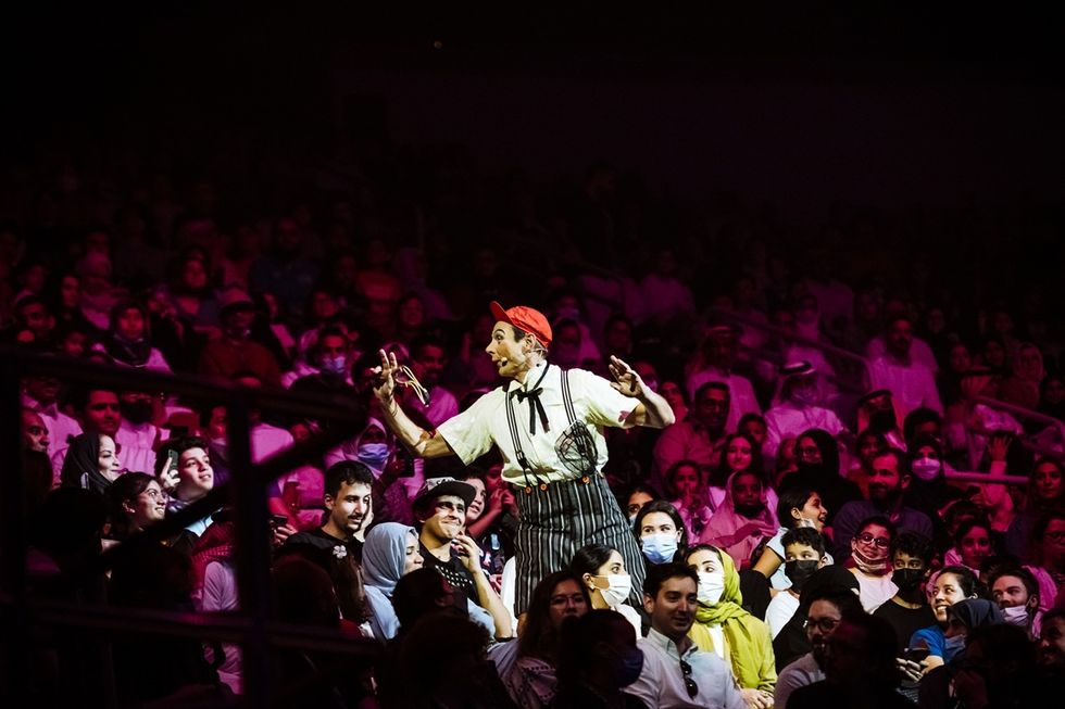 Performer interacts with audience in a crowded theater, under colorful stage lighting in a Cirque du Soleil operformance.